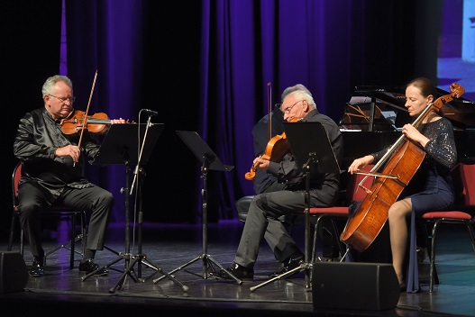 Trójka muzyków, w tym jedna kobieta i dwóch mężczyzn grają na instrumentach smyczkowych na estradzie