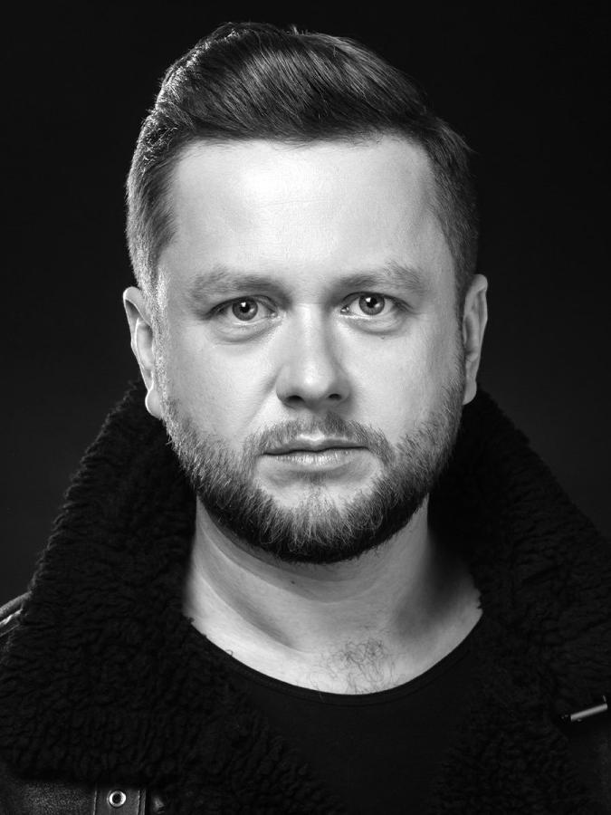 Pianista i aranżer Paweł Tomaszewski ubrany jest w czarną skórzaną kurtkę, posiada lekką bródkę i zaczesane do tyłu włosy