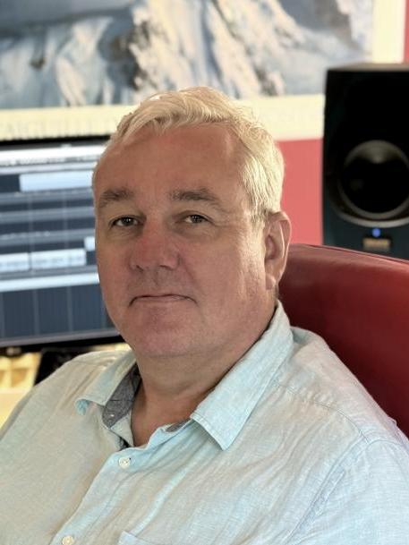 Kompozytor Piotr Majchrzak jest mężczyzną w średnim wieku, siedzi na fotelu w pracowni komputerowej, jest ubrany w białą koszulę