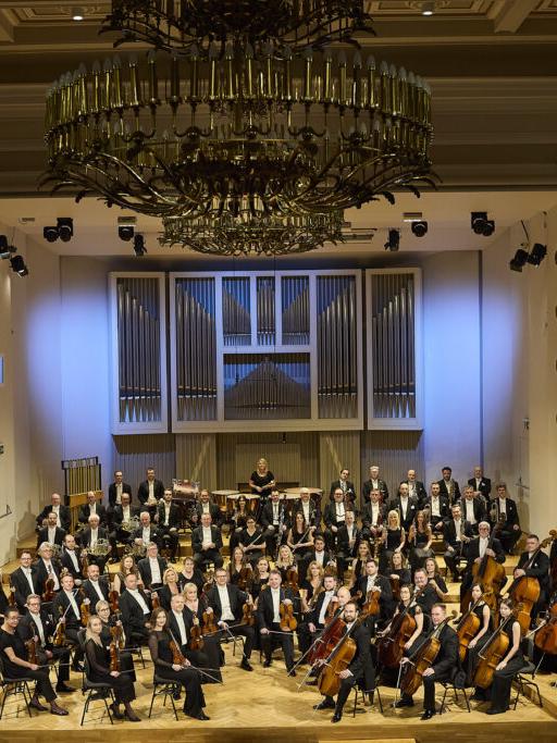 Orkiestra Symfoniczna Filharmonii Śląskiej siedzi na estradzie w składzie około 60 osób, muzycy są ubrani w czarne stroje i trzymają w rękach instrumenty