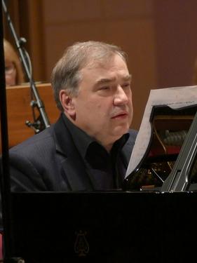 Pianista Marek Mizera siedzi przy czarnym fortepianie, jest ubrany w czarną koszulę, w tle grają muzycy orkiestry