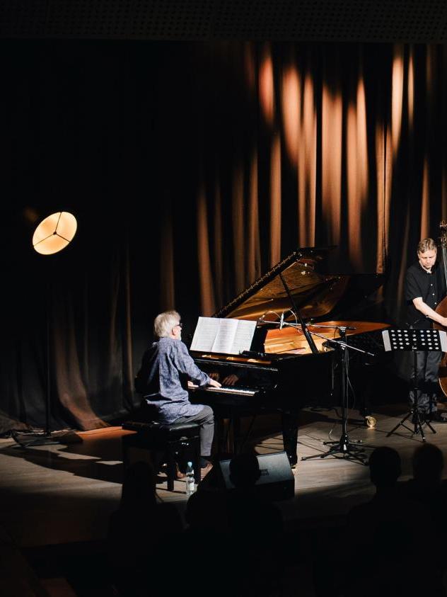 Trio Andrzeja Jagodzińskiego, pianista siedzi przy czarnym fortepianie, kontrabasista stoi z instrumentem, perkusista siedzi za składem instrumentów